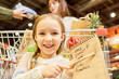 © Seventyfour - Warm-toned portrait of happy little girl grocery shopping in supermarket, smiling looking at camera and pointing at shopping list with food items