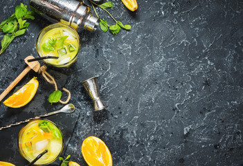 Wall Mural - Cold alcoholic summer citrus cocktail with orange and mint in glasses and on dark stone background. Cocktail making bar tools, shaker, glasses, mint leaves.