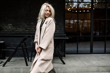 portrait of beautiful young stylish blonde woman wearing beige coat and walking through the city str