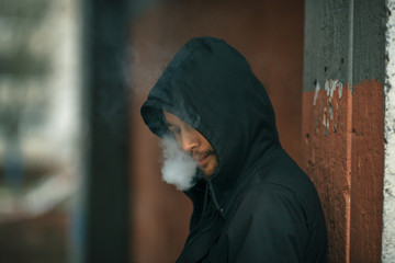 Vape man. Portrait of a bearded handsome young white guy in the hood vaping an electronic cigarette opposite the futuristic urban background.