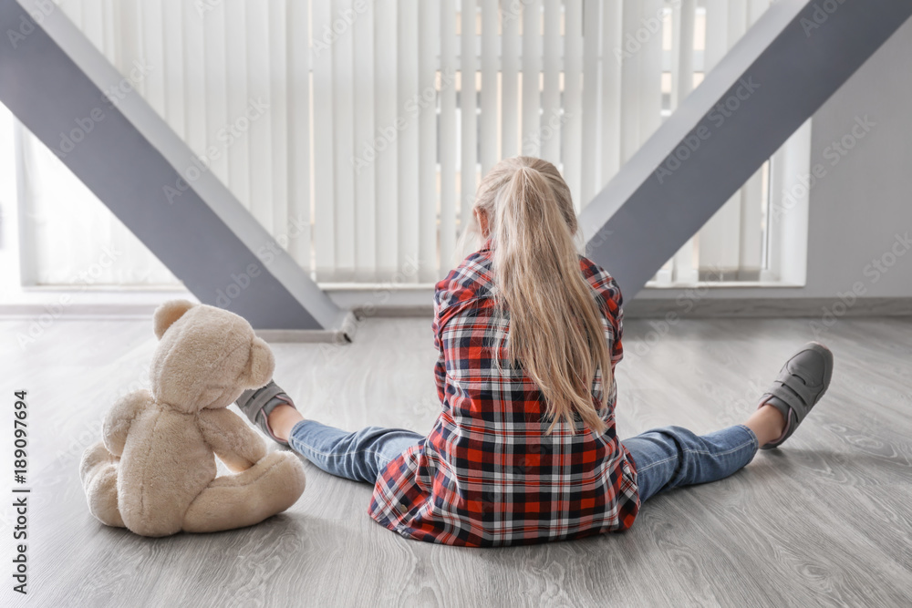 Little girl with teddy bear sitting on floor in empty room. Autism concept