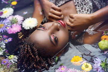 Wall Mural - African woman with bright make up lies in the water with flowers close-up upside-down