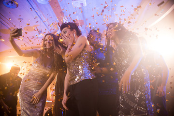 group of beautiful young women wearing glittering dresses dancing under golden confetti and taking s