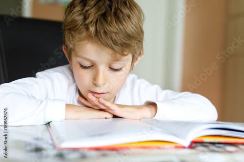 Fototapeta Little blonde school kid boy reading a book at home