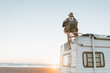 © Alex from the Rock - Charming young woman with hat, poncho,  backpack standing on roof of recreational vehicle on the ocean beach at sunset.