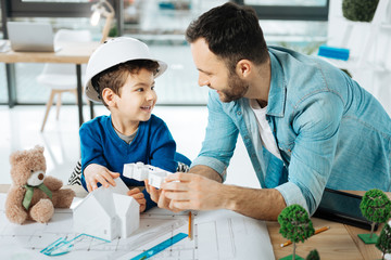 New information. Pleasant young father and his little son in a white hard hat standing in the office near the table and discussing a 3D house model