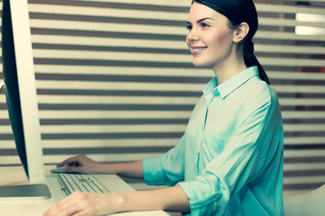 Working with smile. The side view of a pretty dark-haired young woman sitting at the table in the office and working on a computer while smiling