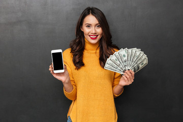 Sticker - Smiling brunette woman holding money and showing blank smartphone screen