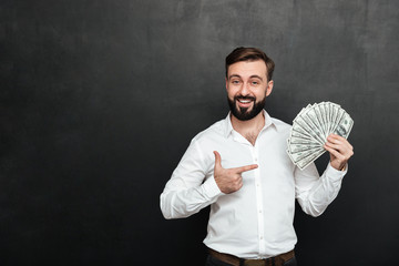 Wall Mural - Portrait of adult man in white shirt posing on camera with fan of 100 dollar bills in hand, being rich and happy over dark gray background