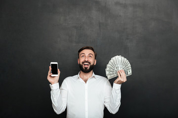 Poster - Portrait of ecstatic man expressing online earnings with holding lots of money dollar currency and smartphone, isolated over dark gray background