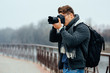 Young stylish photographer holds professional camera, taking photos. Dressed in warm fashionable jacket, scarf, jeans, with black school bag. Outdoors.