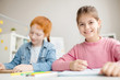 © pressmaster - Cute girl with toothy smile looking at camera at lesson with her classmate near by