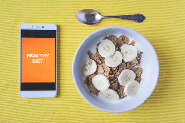 Top view of a white bowl of cereal, orange, lemon, and a mobile phone with written Healthy Diet on the screen on bright yellow background with copy space. Fitness, healthy diet or breakfast concept