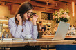 © foxyburrow - Side view. Young business woman is sitting in cafe at table in front of laptop and looks at screen in surprise, lowering her glasses. Hipster girl blogging, learning online. Distance work, education.