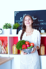 Wall Mural - Smiling young woman holding vegetables standing in kitchen. Smiling young woman