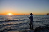 Fototapeta  - silhouette of fisherman with hat on the beach with fish rod standing on sea water fishing at sunset with beautiful orange sky in vacations