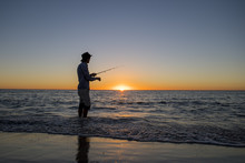 Silhouette Of Fisherman With Hat On The Beach With Fish Rod Standing On Sea Water Fishing At Sunset With Beautiful Orange Sky In Vacations