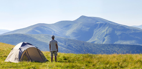 Wall Mural - Hiker man standing near camping tent in carpathian mountains. Tourist enjoy mountain view. Travel concept.