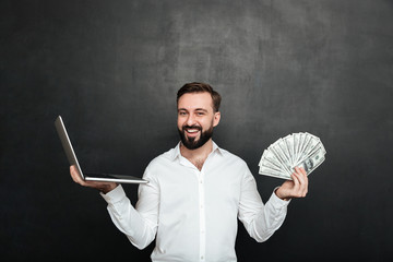 Canvas Print - Portrait of cheerful rich man in white shirt winning lots of money dollar currency using his notebook, over dark gray background