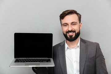Picture of satisfied bearded man holding silver notebook demonstrating or advertising on camera, isolated over gray wall