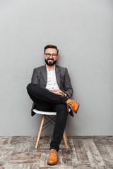 Full-length portrait of relaxed man in casual sitting on chair in office and smiling on camera, isolated over gray background