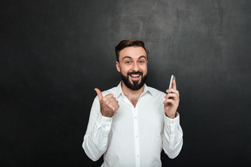 Wall Mural - Satisfied smiling man in white shirt holding smartphone and gesturing thumb aside, over dark gray background