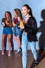 Wall Mural - smiling stylish asian woman holding glass with cocktail while friends drinking cocktails and using smartphone behind at party