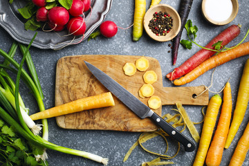 Poster - Chopped carrots on wooden board with kitchen knife and fresh spring vegetables for vegetarian cooking on the table.