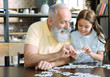 © zinkevych - Let me see. Waist up shot of smart little girl standing next to her granddad and showing him two puzzle pieces while both playing at home.