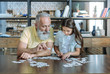 © zinkevych - Smart game. Serene elderly man and his granddaughter sitting at a table and looking at puzzle pieces while playing at home together.