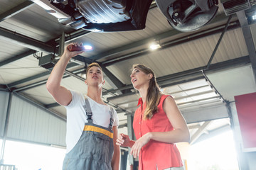 Low-angle view of a reliable auto mechanic, holding a flashlight while checking the parts of the lifted car of a woman in a modern automobile repair shop