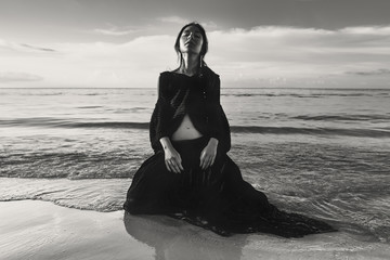 fashionable young woman model sitting on sand in water at sunset