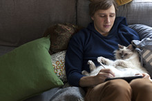 Young Man Sitting With IPad In Sofa With Cat Sleeping On His Belly