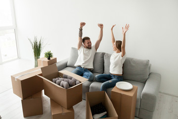 Euphoric young couple feeling happy about moving in new home with boxes, celebrating relocation into own apartment, excited man and woman house owners raising hands proud with buying real estate