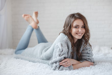 Cute Young Girl In A Gray Knitted Sweater. Beautiful Woman Is Relaxing In A White Bedroom. Beautiful Women In Winter Clothes Are Waking Up In The Morning. Woman Wearing A Sweater In A White Bedroom.