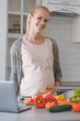 © LIGHTFIELD STUDIOS - smiling pregnant woman standing near vegetables in kitchen