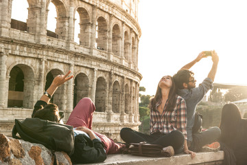 Three young friends tourists sitting lying in front of colosseum in rome taking selfie pictures with smartphone camera. Sunset with lens flare.