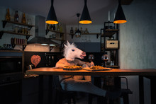 Young Man Sitting At The Table With Food And Drinking Wine In Modern Home Kitchen. Funny Unicorn Dinning Alone At The Bar Counter In Stylish Apartment. Crazy Person. Painted Heart On The Wall