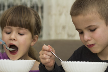 Two children boy and girl eating soup with spoon from a plate with open mouth