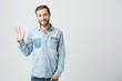 Friendly positive smiling young Caucasian man with stubble and dark hair in trendy denim clothes waving with hand, hailing friends while having fun indoors. Human relations and feelings