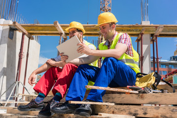 Side view of two workers, reading online information or watching a video on a tablet PC during break at work on the construction site of a contemporary building