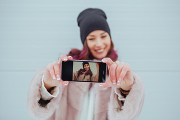 Wall Mural - Fashion portrait of pretty smiling hipster woman in mink coat and smartphone against the colorful grey wall. Making selfie. Casual clothes. Colored hairs