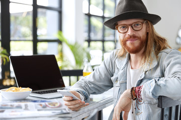 fashionable male with ginger thick beard, wears stylish clothes, reads news in networks, spends time
