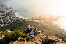 A Couple Is Relaxing On Top Of Lion's Head Mountain In Cape Town And Enjoying The Beautiful Sunset With A View Of Camps Bay And Clifton Beach Areas