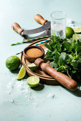 Wall Mural - Ingredients for making mojito cocktail. Bundle of fresh mint, whole and sliced limes, brown sugar, crashed ice cubes, glass of soda water, cocktail tubes on wooden board over green pin up background.