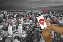 Hands Holding A Phone With Finger Touching The Screen. Red Icon Location Map With City In Background