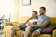 © theartofphoto - Two gay men in casual outfits sitting on sofa embracing and watching TV at home