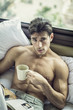 © theartofphoto - Sexy handsome young man laying shirtless on his bed next to window, holding a coffee or tea cup