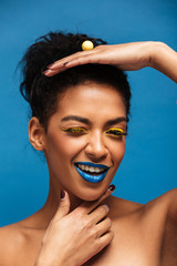 Poster - Vertical image of fancy woman with colorful makeup and curly hair in bun posing on camera with playful look isolated, over blue wall