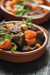 Beef meat stewed with carrots and spices in ceramic pot  on old black wooden board. Selective focus.